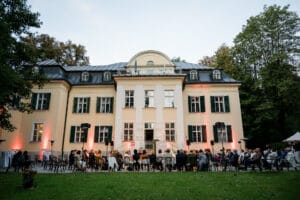 Menschen sitzen draußen und blicken auf ein elegantes, beleuchtetes Herrenhaus mit großen Fenstern und einem angelegten Garten. Der zweistöckige Veranstaltungsort ist perfekt für eine Traumhochzeit. Er verfügt über einen zentralen Bogen und Fensterläden vor einer Kulisse aus Bäumen unter einem klaren Himmel.