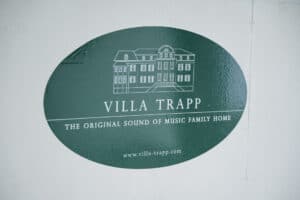 Grünes ovales Schild mit dem weißen Umriss eines großen Hauses. Text: „Villa Trapp, das Original-Familienhaus von Sound of Music“. Dieser Veranstaltungsort ist ideal für Hochzeitsfeiern und bietet eine bezaubernde Kulisse für Ihren besonderen Tag. Website: www.villa-trapp.com.