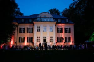 Ein großes, beleuchtetes Gebäude in der Abenddämmerung mit einer Menschenmenge davor, perfekt für eine Hochzeit. Die elegante Fassade verfügt über gewölbte Fenster, die in warmes, rosafarbenes Licht getaucht sind. Hohe Bäume rahmen die Szene ein und schaffen eine ruhige Atmosphäre, ideal für Hochzeiten oder anspruchsvolle Hochzeitsplanung.