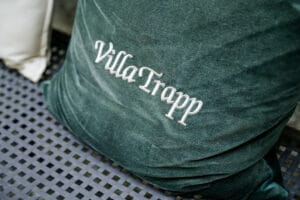 Ein weiches, dunkelgrünes Kissen mit der Aufschrift „Villa Trapp“ in weißer Schrift ruht auf einer strukturierten schwarzen Metalloberfläche und verleiht jeder Hochzeit oder Hochzeitsplanung eine elegante Note.