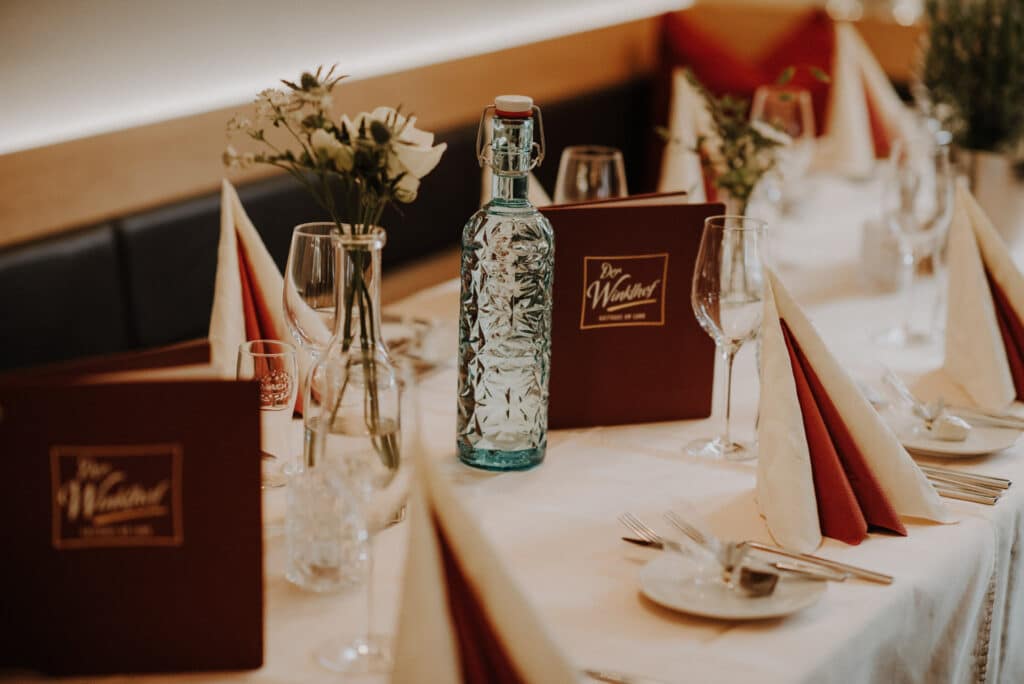 Ein elegant gedeckter Esstisch mit roten und weißen Servietten, Weingläsern und braunen Menüs erweckt den Geist einer anspruchsvollen Hochzeit. Eine dekorative Wasserflasche ziert die Mitte, umgeben von kleinen Blumenarrangements, perfekt für Ihren besonderen Tag.