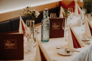 Ein elegant gedeckter Esstisch mit roten und weißen Servietten, Weingläsern und braunen Menüs erweckt den Geist einer anspruchsvollen Hochzeit. Eine dekorative Wasserflasche ziert die Mitte, umgeben von kleinen Blumenarrangements, perfekt für Ihren besonderen Tag.