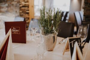 Ein Restauranttisch ist mit leeren Weingläsern, einem silbernen Topf mit Rosmarin, einer roten Speisekarte und einer Namenskarte gedeckt. Perfekt für intime Anlässe und ideal für Hochzeitsplanung. Das Ambiente ist gemütlich, mit Steinwänden und sanfter Beleuchtung.