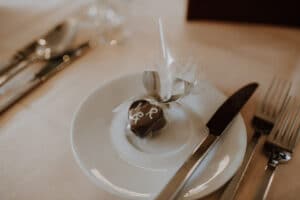 Ein kleines, in braune Schokolade verpacktes Geschenk mit einer weißen dekorativen Schleife wird auf einem weißen Teller platziert, perfekt für jede Hochzeit. Das Besteck, einschließlich Messer und Gabel, wird ordentlich neben dem Teller auf einer hellen Tischdecke arrangiert und verleiht Ihrer Hochzeitsplanung Eleganz.