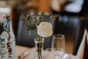 Eine weiße Rose und kleine Wildblumen in einer durchsichtigen Glasflasche auf einem Esstisch, neben einem leeren Weinglas und einer blauen Glasflasche, evozieren den Charme der Hochzeitsplanung. Der sanft verschwommene Hintergrund schafft eine warme, intime Atmosphäre, perfekt für die Feier der Liebe.