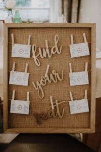 Ein Schild mit einem Holzrahmen und der Aufschrift „Finden Sie Ihren Platz“ in Schreibschrift ziert den Ort der Hochzeit. An Schnüren befestigte Notizkarten zeigen Tischzuweisungen mit Namen und Details vor einem rustikalen Hintergrund aus Sackleinen. Diese elegante Note ergänzt den sanft beleuchteten Innenraum und verleiht Ihrer Hochzeitsfeier Charme.