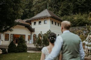 Ein Paar ist von hinten zu sehen, wie es sich umarmt und auf ein charmantes, großes Haus mit Schrägdach blickt, das von üppigem Grün umgeben ist. Diese ruhige Umgebung lässt auf einen intimen Moment schließen, in dem sie von ihrer gemeinsamen Zukunft träumen und vielleicht an Heiraten und Hochzeitsplanung denken.