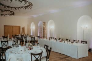 Ein geräumiger, elegant dekorierter Speisesaal, der sich perfekt für eine Hochzeit eignet, mit runden und rechteckigen Tischen mit weißen Tischdecken. Stühle umgeben die mit Kerzen und Blumenarrangements als Tischdekoration geschmückten Tische. Kronleuchter aus Geweihen werten diese exquisite Kulisse für eine unvergessliche Hochzeitsplanung auf.