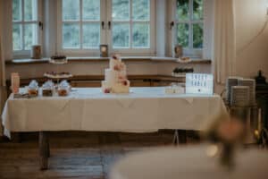Ein Hochzeits-Desserttisch mit einer mehrstöckigen Torte, geschmückt mit rosa Blumen, umgeben von Gläsern mit Bonbons und kleinen Gebäckstücken auf Ständern. Auf einem Schild steht in eleganter Schrift „Sweet Table“, perfekt eingefangen im Schein des natürlichen Lichts. Dieses entzückende Arrangement ist ein wesentliches Element jeder durchdachten Hochzeitsplanung.