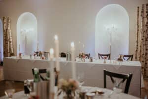 Ein Hochzeitsempfangsraum mit weißen Tischdecken und elegantem Dekor. Auf einem langen Tisch an einer Wand brennen große Kerzen, und die Stühle sind ordentlich angeordnet. Birkenstämme sorgen für eine natürliche Note, während die sanfte Beleuchtung eine warme Atmosphäre schafft – perfekt für alle, die gerade mitten in der Hochzeitsplanung stecken.