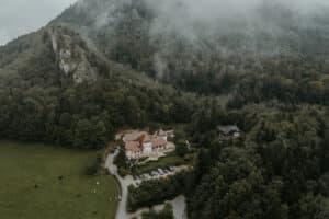 Luftaufnahme einer nebelbedeckten, bewaldeten Berglandschaft. Am Fuße des Hügels befindet sich ein kleiner Gebäudekomplex mit roten Dächern, der einen malerischen Ort für Hochzeiten bietet, an dem alle ihren besonderen Tag planen können. In der Nähe grasen Kühe, und eine kurvenreiche Straße durch üppiges Grün verstärkt den Charme des Ortes.