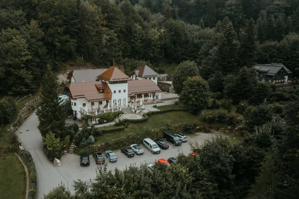 Luftaufnahme eines großen, rustikalen Gebäudes mit rotem Ziegeldach, umgeben von üppigen grünen Bäumen, perfekt zum Heiraten. Vor dem Veranstaltungsort befindet sich ein kleiner Parkplatz für mehrere Autos. Der Veranstaltungsort verfügt über mehrere Ebenen und einen Terrassenbereich, der sich ideal für Hochzeitsfeiern eignet.