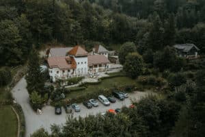 Luftaufnahme eines großen, rustikalen Gebäudes mit rotem Ziegeldach, umgeben von üppigen grünen Bäumen, perfekt zum Heiraten. Vor dem Veranstaltungsort befindet sich ein kleiner Parkplatz für mehrere Autos. Der Veranstaltungsort verfügt über mehrere Ebenen und einen Terrassenbereich, der sich ideal für Hochzeitsfeiern eignet.