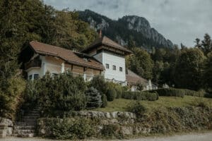 Ein malerisches Gebäude mit Holzbalkon und rotem Dach liegt eingebettet in üppigem Grün am Fuße des Berges unter einem bewölkten Himmel und ist somit ein idealer Ort für eine Hochzeit. Elegant gepflegte Büsche und Bäume umgeben das Anwesen und eignen sich perfekt für alle Hochzeitsplanungsbedürfnisse.