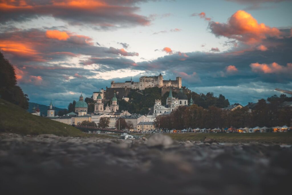 Ein malerischer Blick auf Salzburg, Österreich, mit der Festung Hohensalzburg auf einem Hügel. Wenn der Sonnenuntergang die Wolken dramatisch färbt, kann man sich eine romantische Kulisse vorstellen, die perfekt zum Heiraten ist, inmitten historischer Gebäude und eines charmanten Kopfsteinpflasterwegs.