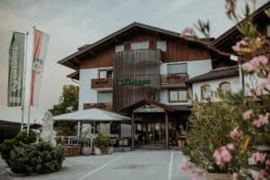 Das Hotel Königgut ist ein rustikales Hotel mit hölzernen Akzenten, Balkonen und Blumenkästen, umgeben von viel Grün und rosa Blumen. Auf der linken Seite stehen Fahnen, und in der Nähe des Eingangs befindet sich eine Außenterrasse mit Sitzgelegenheiten.