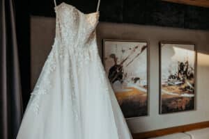 Ein weißes trägerloses Hochzeitskleid mit floralen Spitzendetails hängt im Hotel Königgut vor dem Hintergrund zweier abstrakter Gemälde an der Wand.