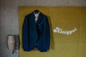 Eine dunkelblaue Anzugsjacke hängt auf einem schwarzen Kleiderbügel vor einer senfgelben Wand mit dem weißen Schriftzug "Königgut", der an den raffinierten Stil des Hotels Königgut erinnert. An der grau gemusterten Wand links ist eine beige Wandlampe angebracht.