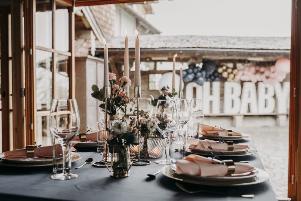 Ein elegant gedeckter Esstisch mit großen brennenden Kerzen, Blumenschmuck und ordentlich gefalteten Servietten auf Tellern lässt auf eine romantische Hochzeitsplanung schließen. Im Hintergrund sind große „OH BABY“-Buchstaben zu sehen, die eine schöne Verbindung von Liebe und Feierlichkeit suggerieren.