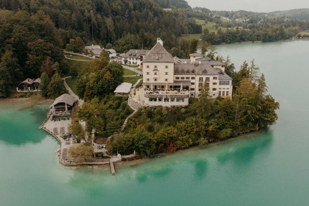 Ein malerisches schlossähnliches Gebäude liegt auf einer üppig grünen Halbinsel, umgeben von türkisfarbenem Wasser, perfekt für eine Traumhochzeit. Die Landschaft ist geprägt von dichten Wäldern und sanften Hügeln, mit anderen kleineren Gebäuden in der Nähe, die eine ruhige, idyllische Umgebung schaffen, die ideal für die Hochzeitsplanung ist.