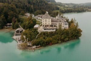 Ein malerisches schlossähnliches Gebäude liegt auf einer üppig grünen Halbinsel, umgeben von türkisfarbenem Wasser, perfekt für eine Traumhochzeit. Die Landschaft ist geprägt von dichten Wäldern und sanften Hügeln, mit anderen kleineren Gebäuden in der Nähe, die eine ruhige, idyllische Umgebung schaffen, die ideal für die Hochzeitsplanung ist.