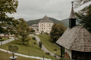 Ein malerischer Blick auf ein historisches Schloss mit steilem Dach, perfekt für eine märchenhafte Hochzeit, eingebettet in eine üppig grüne Landschaft. Eine kleine Kapelle mit Holzdach steht im Vordergrund, während sanfte Hügel die ruhige Szene vervollständigen und sie zu einer idealen Kulisse für eine sorgfältige Hochzeitsplanung machen.