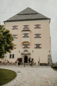Ein hohes, hellgelbes historisches Gebäude mit steilem Dach und roten und weißen Fensterläden bietet eine bezaubernde Kulisse für eine romantische Hochzeit. Zum kunstvollen Eingang führt ein Steinweg. Sitzgelegenheiten im Freien auf einem gepflasterten, von Grün umgebenen Bereich ergänzen den bedeckten Himmel.