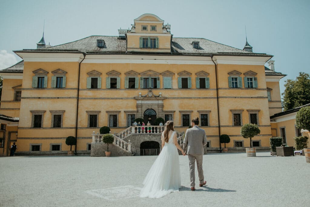 Ein Paar im Hochzeitskleid steht Händchen haltend vor einem großen, eleganten gelben Gebäude, das mit dekorativen Elementen geschmückt ist, und genießt die Freude seiner Hochzeit. Das weiße Kleid der Braut und der graue Anzug des Bräutigams schimmern unter dem klaren, sonnigen Himmel.