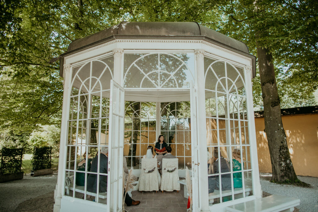 Eine kleine Hochzeitszeremonie findet in einem weißen Pavillon statt, der von üppigen Bäumen umgeben ist. Zwei Personen sitzen einem Geistlichen gegenüber, zu beiden Seiten sitzen einige Gäste. Die Atmosphäre ist intim und ruhig, das Sonnenlicht fällt durch die Blätter – perfekt für diese Hochzeitsmomente.