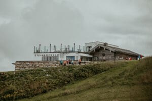 Ein rustikales Gebäude mit der Aufschrift „Steinbockalm“ liegt auf einem grasbewachsenen Hügel unter einem bewölkten Himmel, perfekt für diejenigen, die ihre Hochzeit planen. Eine Gruppe von Menschen steht draußen in der Nähe des Eingangs, mit einer Kulisse aus Kiefern und Glasgeländern.