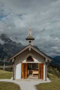 Eine kleine Kapelle mit Holztüren und einem Kirchturm mit Kreuzspitze steht auf einem grasbewachsenen Hügel und ist perfekt für eine intime Hochzeit. Berge und Wolken bilden eine dramatische Kulisse, gedämpftes Licht sorgt für eine ruhige Atmosphäre. Das Innere der Kapelle, ideal für Hochzeitsplanung, ist durch offene Türen sichtbar.