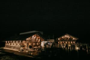 Ein gemütliches Alpenchalet ist warm beleuchtet unter einem sternenklaren Nachthimmel, perfekt für eine romantische Hochzeit. Holzarchitektur und Sitzgelegenheiten im Freien schaffen eine rustikale Atmosphäre, ideal für diejenigen, die heiraten möchten. Dezente Lichter akzentuieren das Gebäude und werfen einen sanften Schein auf die bezaubernde Umgebung.