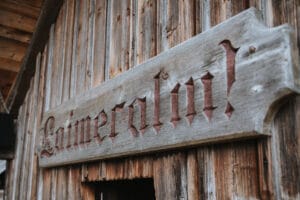 Ein verwittertes Holzschild mit der Aufschrift „Laimeralm“ in rustikalen, dunkelroten Buchstaben ist an der Außenseite eines alten Holzgebäudes angebracht, das sich perfekt für eine Hochzeit eignet. Der Veranstaltungsort mit seinen vertikalen Brettern und dem schrägen Dach bietet eine bezaubernde Kulisse für diejenigen, die von einer Hochzeit inmitten der Natur träumen.