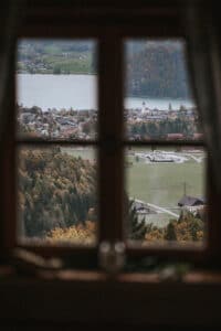 Blick durch ein Fenster auf eine malerische Landschaft mit einem See, einer kleinen Stadt und umgebenden bewaldeten Hügeln unter einem bewölkten Himmel. Die heitere Schönheit ist perfekt für die Planung Ihrer Traumhochzeit in der Umarmung der Natur in der malerischen, am Wasser gelegenen Stadt.