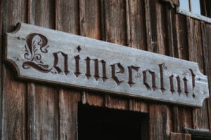 Ein rustikales Holzschild mit kunstvoller gotischer Schrift hängt an einer alten Holzwand. Der Text, der wahrscheinlich „Laimecalnt“ lautet, lässt auf einen historischen Charme schließen, perfekt für eine intime Hochzeit oder eine durchdachte Hochzeitsplanung in einem zeitlosen Ambiente.