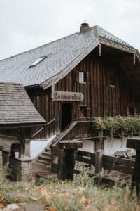 Eine rustikale Holzhütte mit grauem Schindeldach steht mitten im Grünen und eignet sich perfekt für eine intime Hochzeitsplanung. Über dem Eingang steht ein Schild mit der Aufschrift „Kaineralm“. Holzgeländer und ein Steinfundament vervollständigen die traditionelle alpine Architektur, ideal für diejenigen, die in der Natur heiraten möchten.