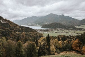 Eine malerische Landschaft, ideal für eine Traumhochzeit, mit einem Tal mit einem charmanten Dorf, umgeben von üppigen, herbstlich gefärbten Wäldern. Im Hintergrund schaffen teilweise in Wolken gehüllte Berge eine romantische Atmosphäre, perfekt für eine durchdachte Hochzeitsplanung unter einem bedeckten Himmel.