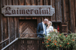 Ein Paar küsst sich während seiner Hochzeit auf einem rustikalen Holzbalkon. Der Mann trägt einen Anzug und die Frau hält einen Strauß Sonnenblumen in der Hand. Über der Tür steht ein Holzschild mit der Aufschrift „Laimeralm“. Leuchtend orange Blumen in einem Blumentopf verleihen diesem bezaubernden Moment einen Farbtupfer.