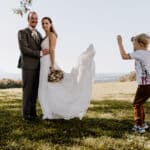 Ein Paar im Hochzeitskleid posiert an einem sonnigen Tag im Freien für ein Foto und fängt die Essenz ihrer perfekten Hochzeit ein. Die Braut hält einen Blumenstrauß, während der Bräutigam einen Anzug trägt. Ein kleiner Junge, leger gekleidet, springt verspielt in der Nähe vor einer Kulisse aus Bäumen und einer entfernten Landschaft herum.