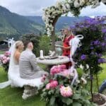 Bei einer bezaubernden Hochzeit im Freien sitzt ein Paar unter einem mit weißen und violetten Blumen geschmückten Blumenbogen. Eine Frau in einem roten Kleid steht und führt die Trauung durch. Die Kulisse aus üppigem Grün und fernen Bergen rundet diese perfekte Szene für alle ab, die in Heiratsstimmung sind.