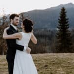 Braut und Bräutigam umarmen sich im Freien mit einer atemberaubenden Berglandschaft im Hintergrund und feiern ihre Hochzeit. Die Braut trägt ein atemberaubendes weißes Kleid und der Bräutigam einen flotten dunklen Anzug. Sie lächeln sich liebevoll an, umgeben von der Schönheit der Natur.