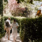 Ein Paar geht Händchen haltend einen sonnenbeschienenen Kiesweg entlang, der von üppigen grünen Hecken umgeben ist. Die Frau im weißen Kleid und der Mann in Weste und heller Hose strahlen Freude aus, während sie ihre Hochzeitspläne besprechen. Sonnenlicht fällt durch die Bäume und schafft eine friedliche und romantische Atmosphäre.