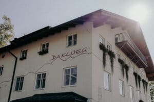 Das beigefarbene Gebäude mit den dunklen Holzschindeln, das den Namen „DAKLUEG“ trägt, strahlt eine bezaubernde Atmosphäre aus, die sich perfekt für Hochzeitsfeiern eignet. Mehrere mit Blumenkästen geschmückte Fenster fangen das Sonnenlicht ein, während Bäume im Hintergrund eine natürliche Note verleihen.