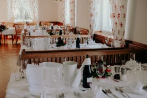 Ein Speisesaal mit Holzstühlen und -tischen, elegant gedeckt für eine Hochzeitsveranstaltung, mit weißen Tischdecken, Gläsern, Besteck und Weinflaschen. Blumenarrangements und natürliches Licht durch große Fenster werten das Dekor auf, perfekt für eine durchdachte Hochzeitsplanung.