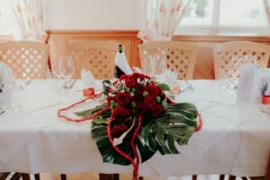 Ein wunderschön gedeckter Esstisch für eine Hochzeit, geschmückt mit einem Strauß roter Rosen und Grünpflanzen als Tafelaufsatz. Das Gedeck besteht aus Weingläsern, Servietten und einer in ein weißes Tuch gewickelten Flasche. Im Hintergrund stehen Holzstühle neben einem Fenster mit hellen Vorhängen. Perfekt für Ihre Hochzeitsplanung.