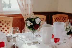 Ein für eine Hochzeit gedeckter Esstisch mit Weingläsern, Salz- und Pfefferstreuern, einem kleinen Blumengesteck aus roten und weißen Blüten und weißen Eimern. Der Tisch ist mit einer weißen Tischdecke bedeckt und steht neben einem Fenster mit elegant drapierten Vorhängen.