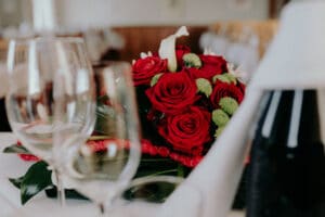 Ein Strauß roter Rosen mit grünen Akzenten ruht auf einer weißen Tischdecke und bereitet die Bühne für eine unvergessliche Hochzeit. Leere Weingläser umgeben das Arrangement und sorgen für eine elegante und romantische Atmosphäre, die perfekt für jede Hochzeitsplanung ist.