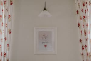 Ein gerahmtes Kunstwerk mit den Worten „Unsere Liebe für immer“ und einem roten Herz hängt an einer beigen Wand und verleiht dem Raum eine romantische Note, die perfekt für alle ist, die ihre Hochzeit planen. Darüber erhellt eine Lampe mit weißem Schirm den Raum, während Vorhänge mit roten Blumenmustern eine charmante Note verleihen.