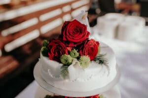 Eine Hochzeitstorte, geschmückt mit leuchtend roten Rosen und kleinen grünen Blumen, ist eine anmutige Ergänzung für den besonderen Tag des Paares. Der weiße Zuckerguss der Torte bietet einen klaren Hintergrund, an den Rändern sind zarte dekorative Details sichtbar – ein exquisiter Mittelpunkt für jede Hochzeitsfeier.
