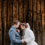 Braut und Bräutigam stehen Stirn an Stirn, halten sich an den Händen und lächeln glücklich an ihrem Hochzeitstag. Die Braut trägt ein weißes Kleid und einen Blumenkopfschmuck, während der Bräutigam einen grauen Anzug mit einer roten Ansteckblume trägt. Sie stehen vor einer rustikalen Holzwand und fangen die Essenz perfekter Hochzeitsplanung ein.